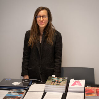 A woman in a black blazer stands smiling behind a table stacked with various books.