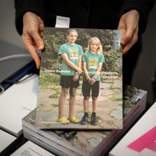 A light skinned person holds a book to the camera. Its cover depicts two white skinned children wearing green tee shirts that say "Brother For Sale $1" and "Sister For Sale $1". The book is titled "Clara Gutsche: Portraits d'enfants"