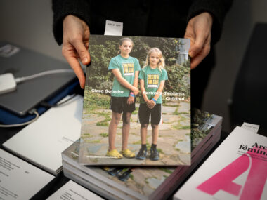 A light skinned person holds a book to the camera. Its cover depicts two white skinned children wearing green tee shirts that say "Brother For Sale $1" and "Sister For Sale $1". The book is titled "Clara Gutsche: Portraits d'enfants"