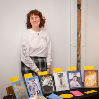 A women in a white, long sleeve shirt stands behind a table displaying risograph printed zines.