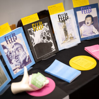 A table with a black tablecloth displays brightly coloured editions of FEELS zine. A mannequin hand holds green business cards.