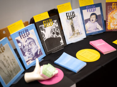 A table with a black tablecloth displays brightly coloured editions of FEELS zine. A mannequin hand holds green business cards.