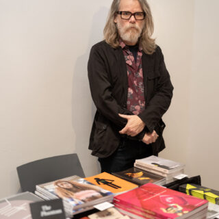 A bearded man in a suit jacket and colourfully patterned shirt stands behind a table stacked with books.