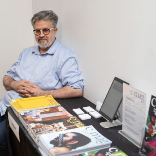 A man in a light blue collared shirt sits beside a table displaying multiple editions of C Magazine.