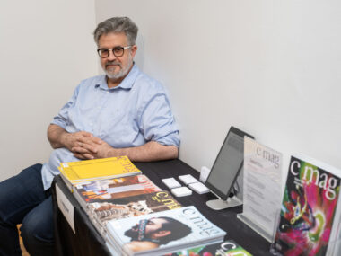 A man in a light blue collared shirt sits beside a table displaying multiple editions of C Magazine.