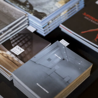 A stack of art exhibition catalogs sit on a table with a black table cloth