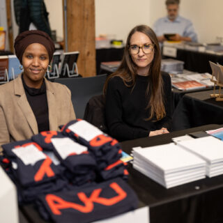 A dark skinned woman and a light skinned woman sit smiling behind a table displaying T-shirts and multiple copies of a white covered book.