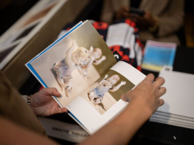 A dark skinned hand holds a book showing a two page spread depicting a sculpture of dolls marked with black insect silhouettes.