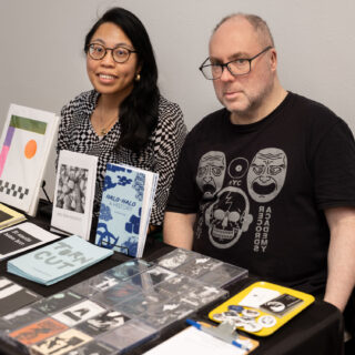 A white man and an Asian woman sit behind a table with cassette tapes, zines, and prints on display.