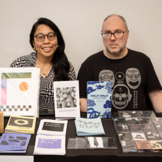 A white man and an Asian woman sit behind a table with cassette tapes, zines, and prints on display.