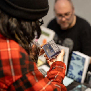 A person with dark brown hair, wearing a red flannel and black beanie examines two cassette tapes.