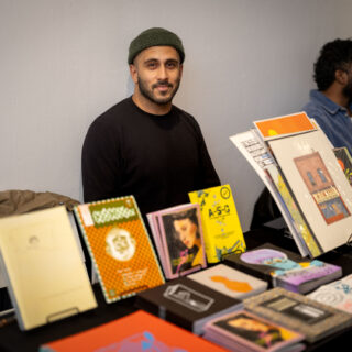 A brown-skinned man wearing a green beanie and black shirt sits smiling behind a table with various, colourful prints and zines on display.