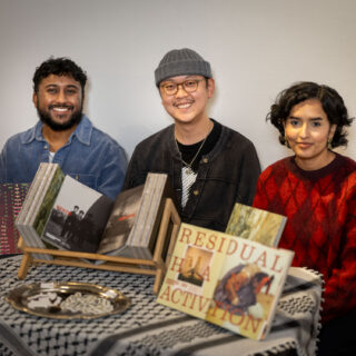 Three smiling, multi racial people sit behind a table with PATINAPATINA Magazine on display. A kuffiyeh is draped on the table.