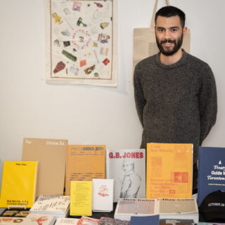 A smiling man with a beard stands behind a table full of various publications from Art Metropole.