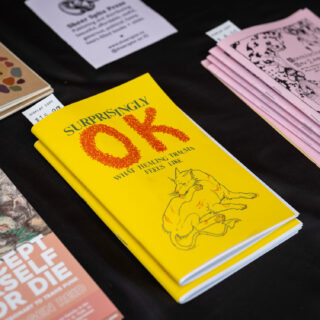 On a black background, brightly coloured zines sit on display. The focal point is a bright yellow booklet titled "Surprinsly OK", with an illustration of an injured dog licking its wounds on the cover.