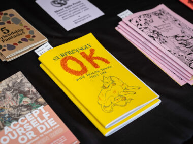 On a black background, brightly coloured zines sit on display. The focal point is a bright yellow booklet titled "Surprinsly OK", with an illustration of an injured dog licking its wounds on the cover.