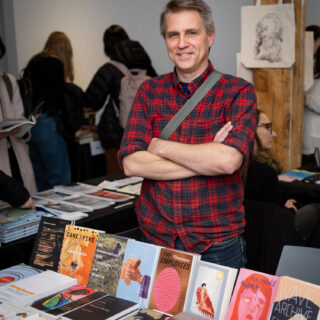 A smiling, white man wearing a red plaid shirt stands behind a table with various publications from Book*Hug Press.