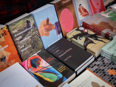 A table stacked full with multiple books, including titles "My Conversations with Canadians", "Dry Your Tears to Perfect your Aim", and "Before I Was a Critic, I Was a Human Being".