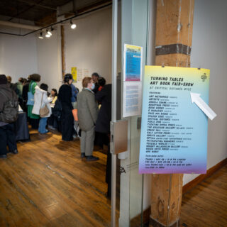 A blue, purple, and yellow gradient sign reading "Turning Tables Art Book Fair" point with a white arrow into a crowded gallery space with wooden floors.