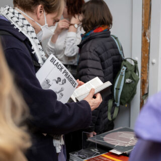 A light skinned person wearing a kuffiyeh scarf and an N95 face mask holds a copy of a magazine with the words "Black Prairies" while leafing through another publication.