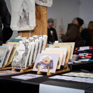 A black table-clothed table displays multicoloured illustrative prints by Angela Zhang on bamboo racks.