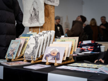 A black table-clothed table displays multicoloured illustrative prints by Angela Zhang on bamboo racks.