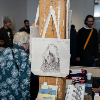 A tote bag with a black and white illustration of a house hangs on a wooden pillar in a busy gallery.