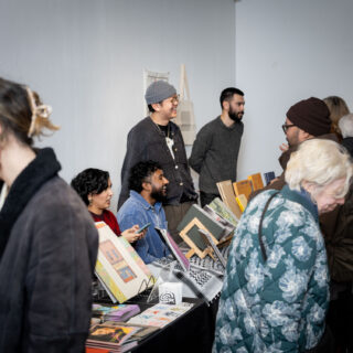 A diverse crowd of smiling people of various skin tones and ages mingle around tables displaying books, zines, and other art materials.