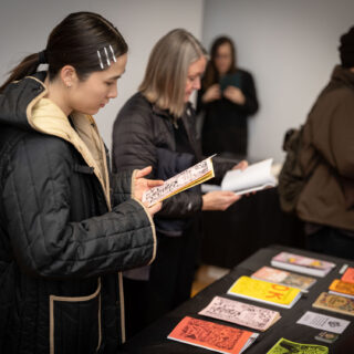 A person in a black and beige winter jacket looks thoughtfully at a zine in front of a table displaying multicoloured zines by Sheer Spite Press.