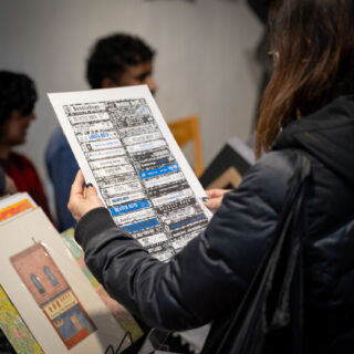 A light skinned person in a black jacket holds a large, white poster of Beastie Boys cassette tapes.