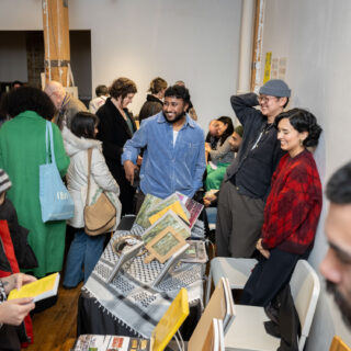 Three multi-racial, smiling people stand behind a table with a kaffiyeh, displaying magazines for sale while a small crowd walks by them.