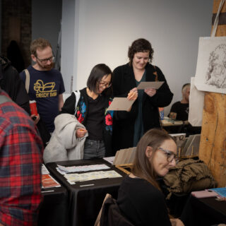 A group of three people look at prints in front of a black -table clothed table displaying zines and prints.