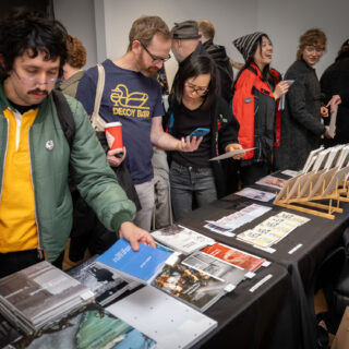 A spectacled man with a moustache browses through a table of books. He is holding a blue, softcover book, and wearing a yellow hoodie with a green puff jacket and backpack.