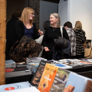 Two smiling, chatting women stand behind a black table-clothed table with books stacked.