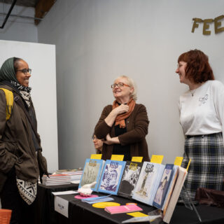 Three women of different races stand smiling in front of a table-clothed table displayed with risograph printed zines.