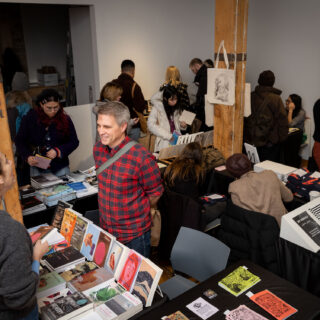 A Birds Eye view photo shows a crowded gallery space with multiple black table-clothed tables showcasing a variety of books and zines. Smiling crowds linger around browsing.