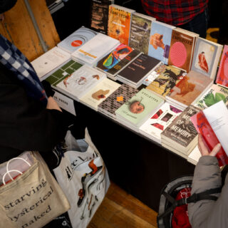 A table with a black table cloth is filled with multiple books from Book Hug Press, two people browse the selection.