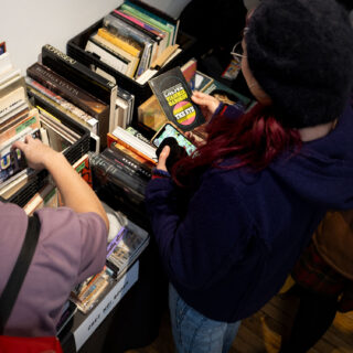 A feminine person with dyed red hair and a purple sweater looks at a vintage copy of "The Eye" by Vladimir Nabokov while another person browses through other vintage books displayed beside them.