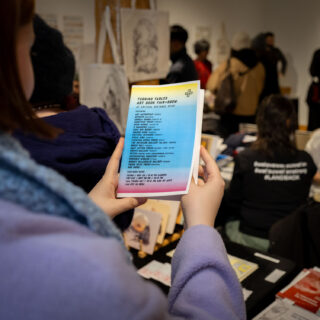 A woman in a light purple jacket holds up a colourful flyer reading "Turning Tables Art Book Fair" with a list of vendors.