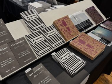 A white display rack sits on a table with a black tablecloth, multiple titles are visible including "Property Journal", with a black and white striped cover, and "Saborami" with a beige and red cover.