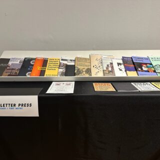 A table with a white display rack and a black tablecloth showcases various zines.