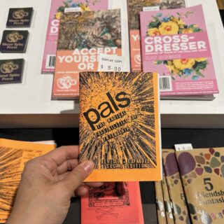 A light-brown skinned hand holds a bright orange coloured zine with a black illustration titled "pals".