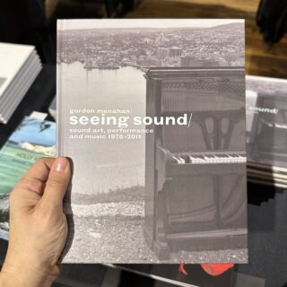 A light skinned hand holds a grey toned art book titled "Gordon Monahan / Seeing Sound"