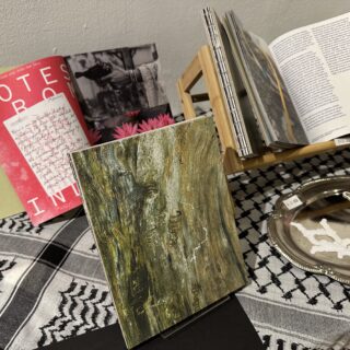 A table with a kuffiyeh table cloth displays multiple copies of a hardbound, art publication. The cover is a green, wood patterned photograph with termite scripture.