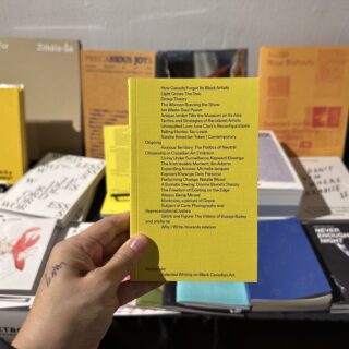 A light skinned hand holds a bright yellow, softcover book titled "Selected Writings on Black Canadian Art" in sans serif black text.