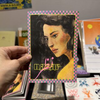 A light skinned hand holds a zine titled "Nazar", the cover depicts a risograph printed drawing of a young woman with long black hair, framed by a pink and blue pattern.