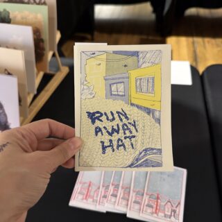 A light skinned hand holds a small, risograph printed zine titled "Run Away Hat".