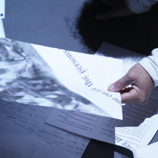 A hand holds a sheet of paper with distorted text moving diagonally across its surface, surrounded by more scattered papers underneath it, illuminated with a soft light from above.