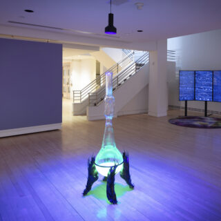 An art installation of a glowing green glass object lit with purple light, surrounded by dark and abstract sculptural elements at its base, accompanied by a digital display in the background comprise of three flatscreen monitors.