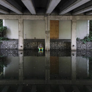 Under an overpass, and surrounded by still water, greenery, and concrete columns, a figure in the distance is holding a green object and leaning down to the water’s edge.